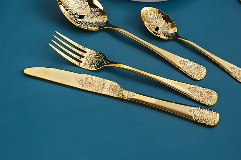 Stainless steel gold flatware set by MC THE BLACK DIAMOND, includes knives, forks, and spoons arranged on a table.